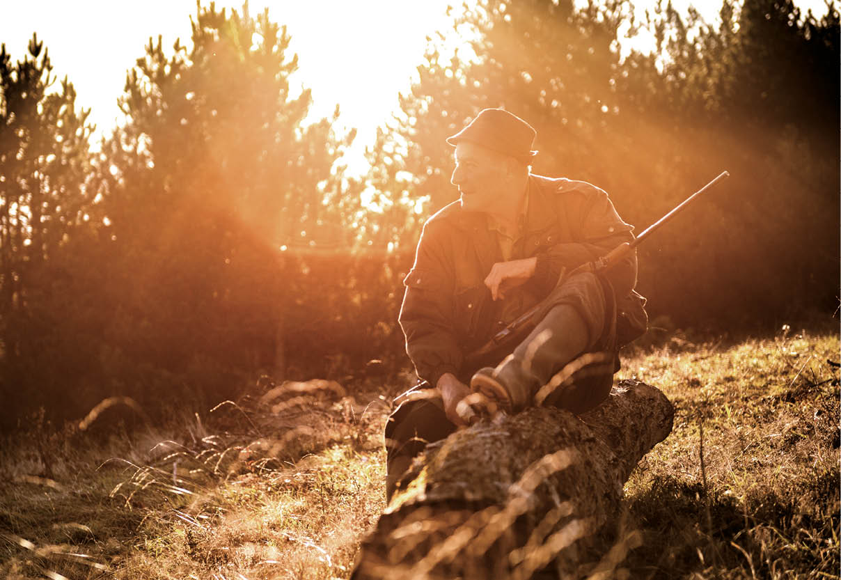 Portrait of a hunter in the forest while sitting on the tree log and holding a rifle.
