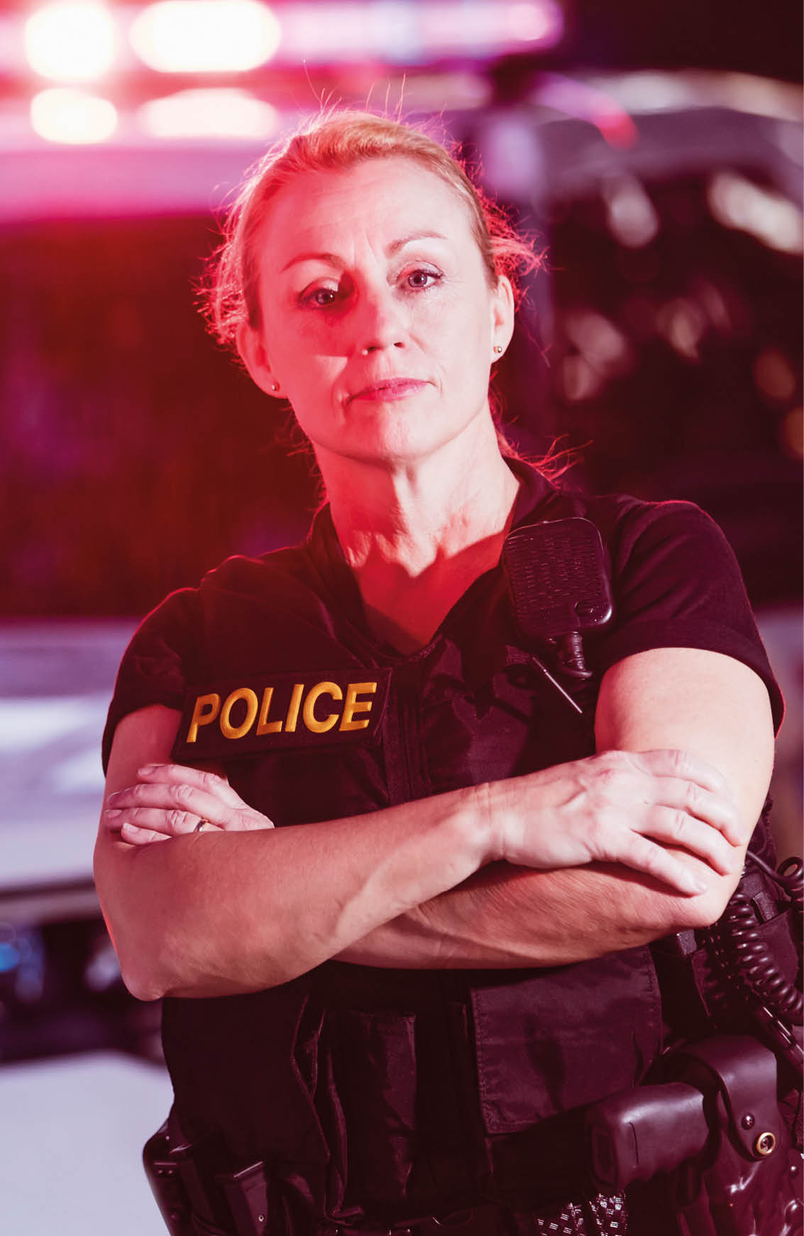 A policewoman wearing a bulletproof vest, standing beside police cars with emergency lights flashing.
